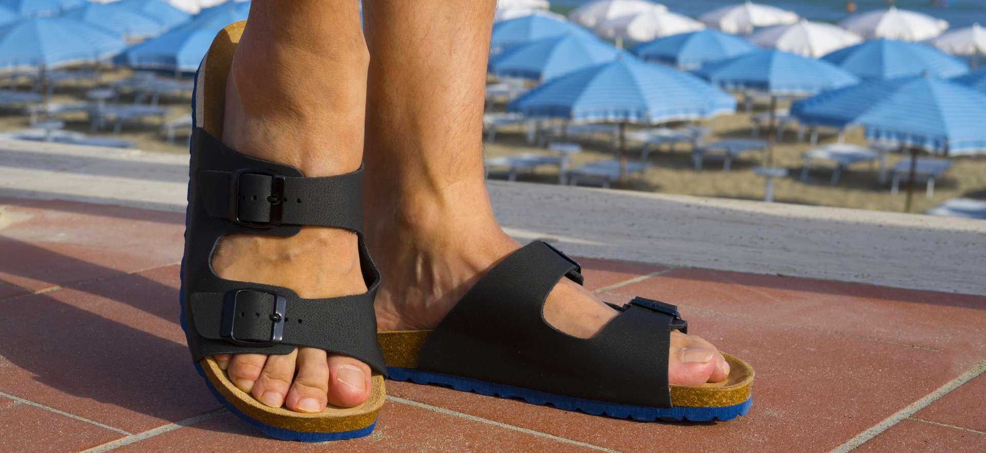Osoba nosi crne natikače s kopčama na pločniku ispred plaže s plavim suncobranima i ležaljkama.