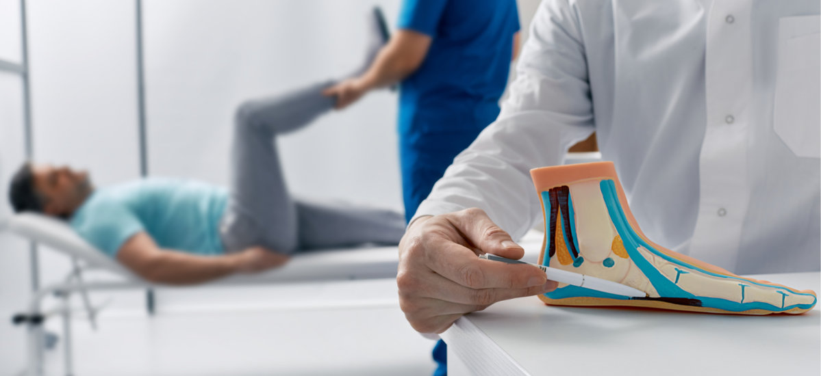Ordinacija, osoba leži na krevetu dok druga drži anatomski model stopala. Na slici se nalaze dvije osobe u ordinaciji. Jedna osoba leži na krevetu, dok druga drži anatomski model stopala u ruci.